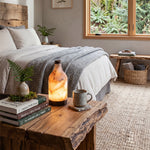 Aldric Alabaster Flask Table Lamp | Natural Alabaster Stone and Bronze Metal | Illuminated on a live-edge wooden bench in a cozy rustic bedroom with neutral bedding | Severn Lighting