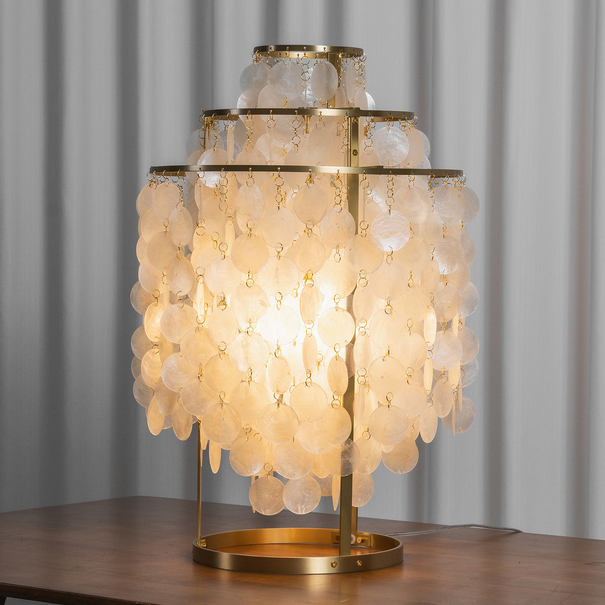 Callista Tiered Capiz Table Lamp | Capiz, Metal | Close-up staged shot of the fully illuminated capiz shell table lamp casting a heavy golden glow onto a polished dark wood table surface | Severn Lighting