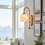 Cordelia Alabaster Globe Wall Sconce | Natural Alabaster and Brushed Brass | Mounted on a white wall by a window overlooking a beautiful coastal city skyline view | Severn Lighting
