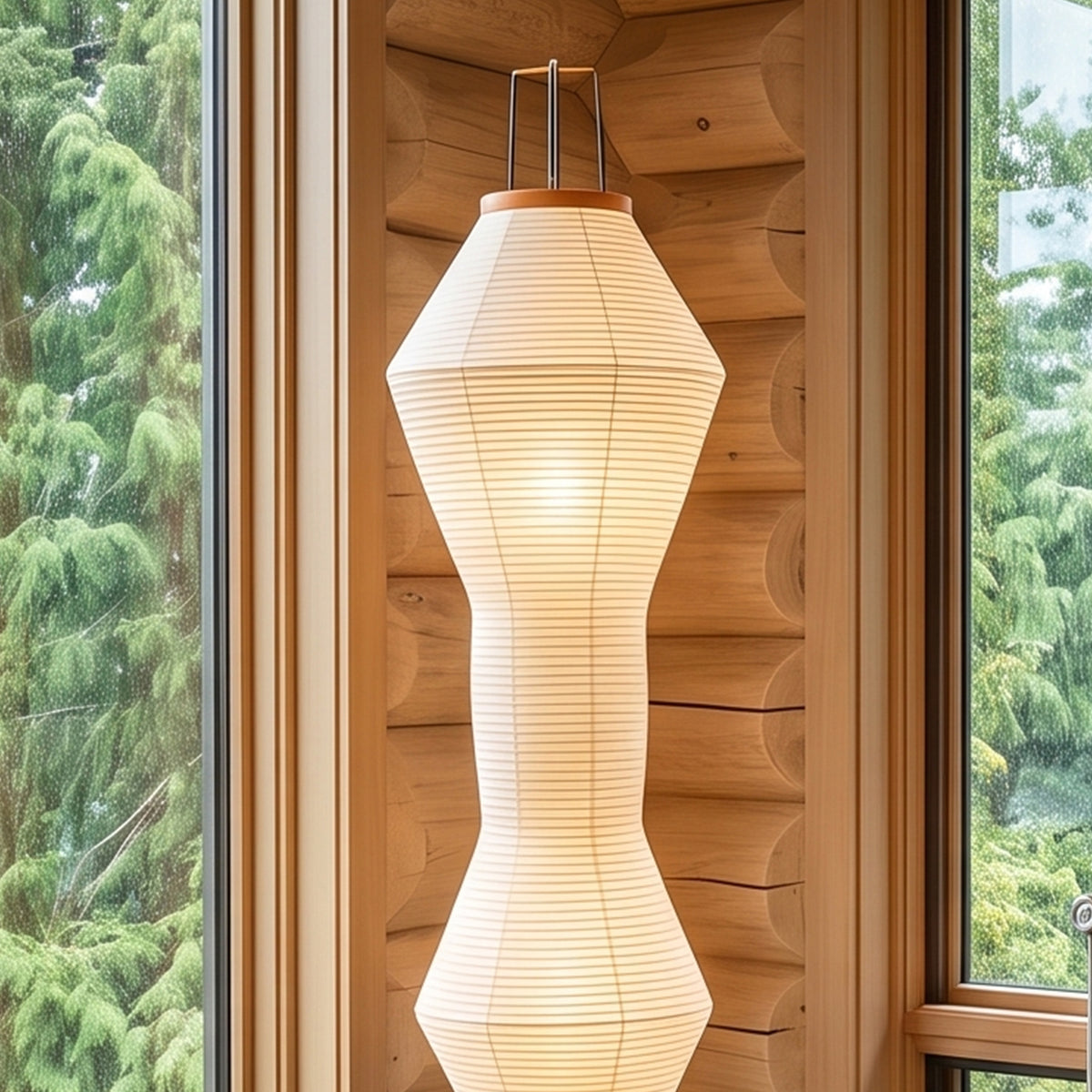 Cosima Hourglass Washi Paper Floor Lamp | Japanese Wabi-Sabi Paper and Black Tripod | Vertical shot of the glowing hourglass paper shade near a log cabin wall window | Severn Lighting