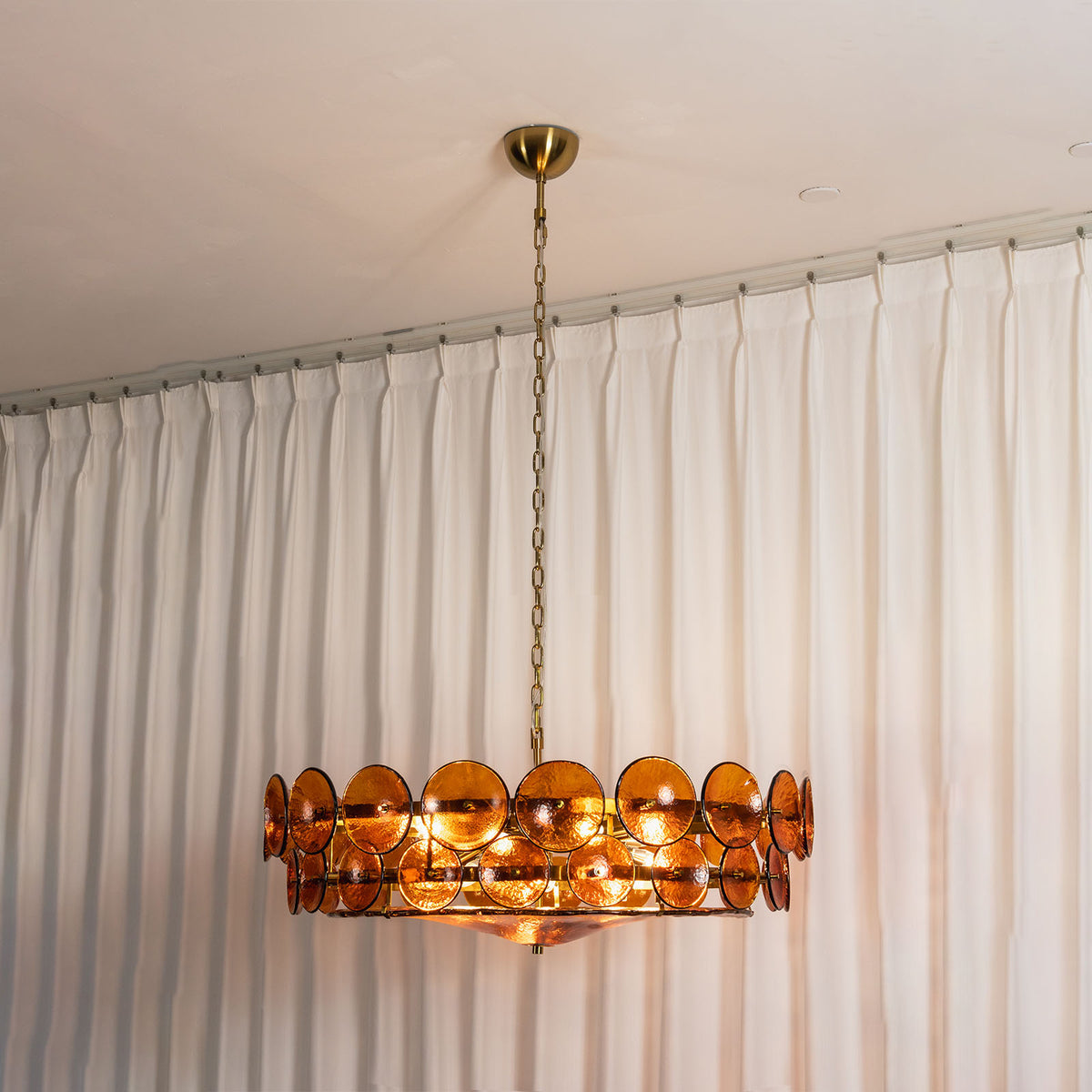 Lovenne Textured Glass Disc Ring Chandelier | Textured Glass and Brass | Real-life view of amber glass variant glowing softly near a white curtain | Severn Lighting