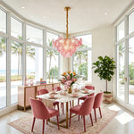 Rosalind Petal Glass Chandelier | Textured Spiral Pink Glass and Gold Metal | Hanging in a luxurious high-ceiling dining room above a pink chair setup by large windows | Severn Lighting