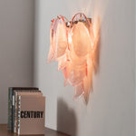 Rosalind Rose Glass Wall Sconce | Glass, Metal | Side angle view of the illuminated pink glass petal fixture casting a cozy light halo on a wall near decorative books | Severn Lighting