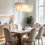 Sirena Capiz Elongated Linear Chandelier | Capiz Shell, Metal | Positioned above a sleek marble dining table with upholstered dining chairs next to a large window | Samu Lamp Sirena Capiz