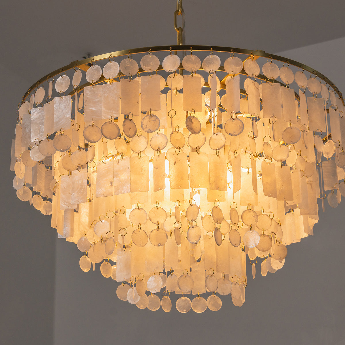 Thalassa Tiered Capiz Shell Chandelier | Capiz Shell, Metal | Angular close-up of the fully illuminated tiered chandelier radiating an intense, warm golden ambient glow from within the shell layers | Severn Lighting