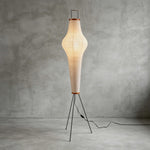 Verena Ribbed Paper Column Floor Lamp | Ribbed Paper and Metal Tripod Legs | Full studio display against a rustic textured grey wall background with the column lamp fully lit | Severn Lighting