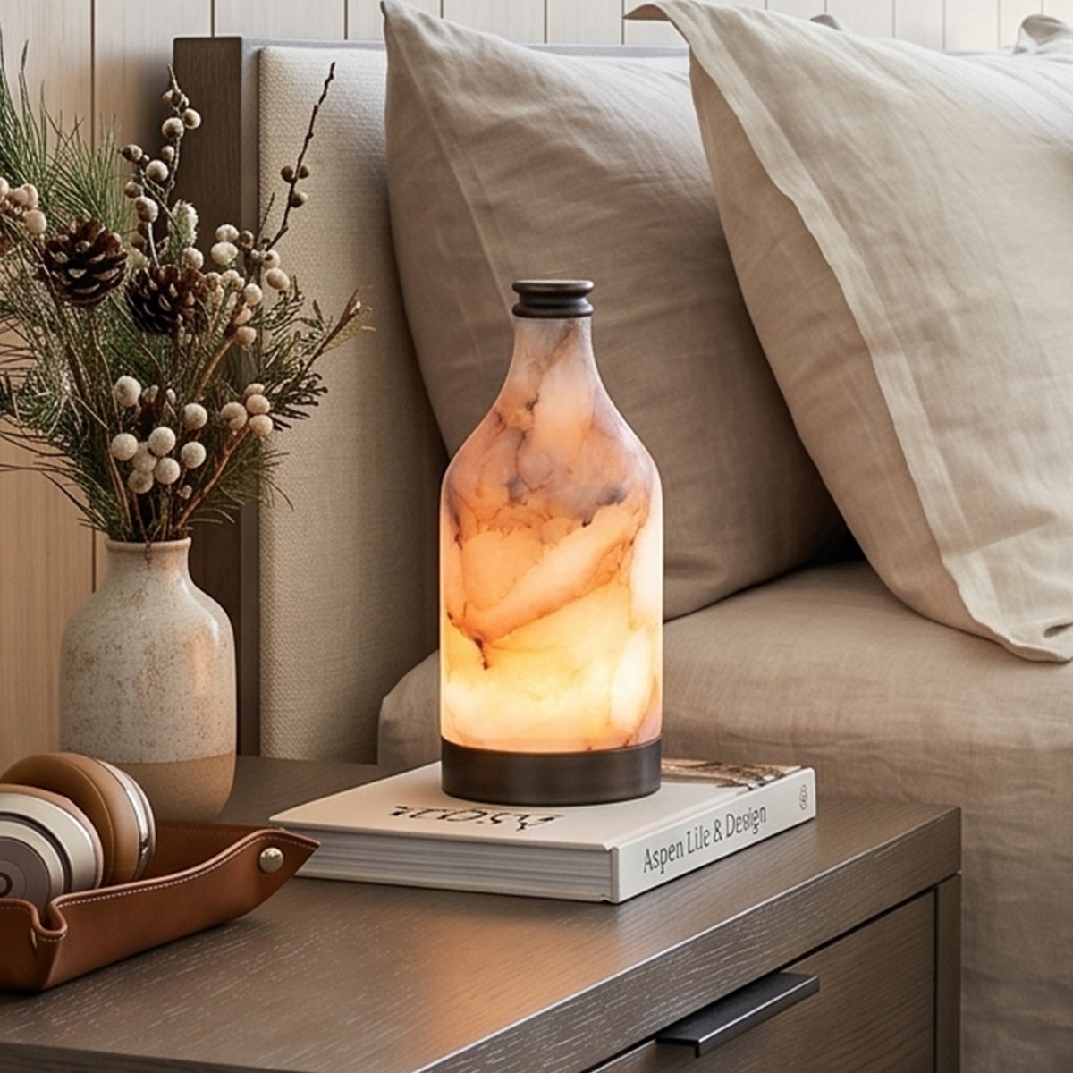 Aldric Alabaster Flask Table Lamp | Natural Alabaster Stone and Bronze Metal | Close-up scene of the lamp glowing softly on a side table next to beige linen pillows and dry flowers | Severn Lighting