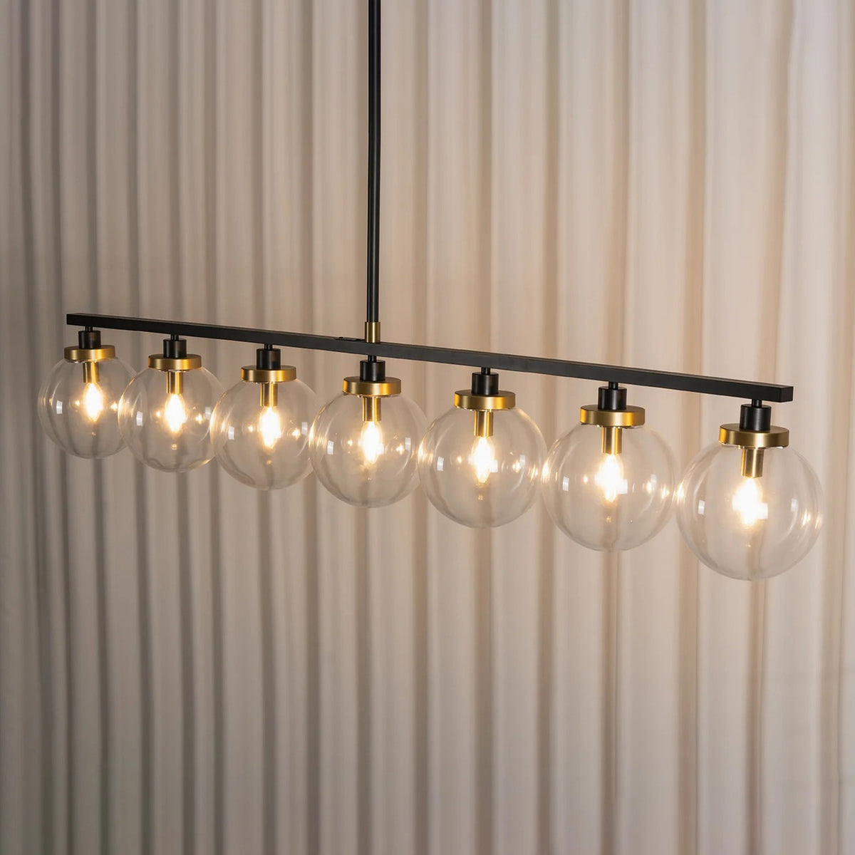 Alistair Linear Glass Globe Chandelier | Hanging Dining Room Lamp | Studio side perspective view of the 8-light linear suspension fixture illuminated against white pleated drapes | Severn Lighting