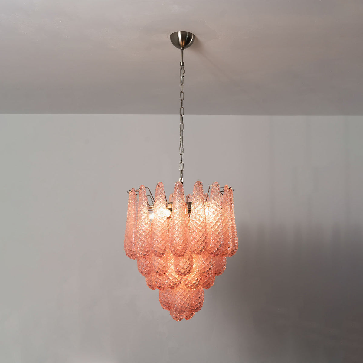 Aubine Pink Textured Glass Tiered Chandelier | Pink Textured Glass and Brass | Full view glowing softly displayed on a neutral studio wall | Severn Lighting