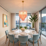 Aubine Pink Textured Glass Tiered Chandelier | Pink Textured Glass and Brass | Hanging above a round marble dining table with blue chairs and ocean views | Severn Lighting