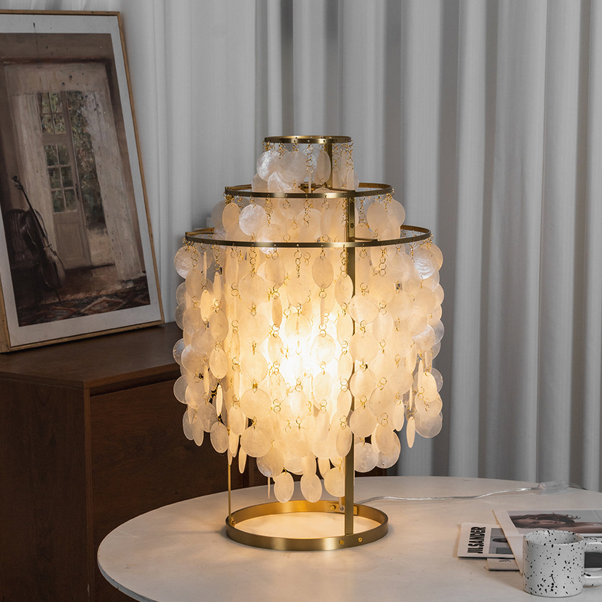 Callista Tiered Capiz Table Lamp | Capiz, Metal | Fully lit capiz shell table fixture radiating a cozy warm glow on a round white table in front of light grey drapes | Severn Lighting