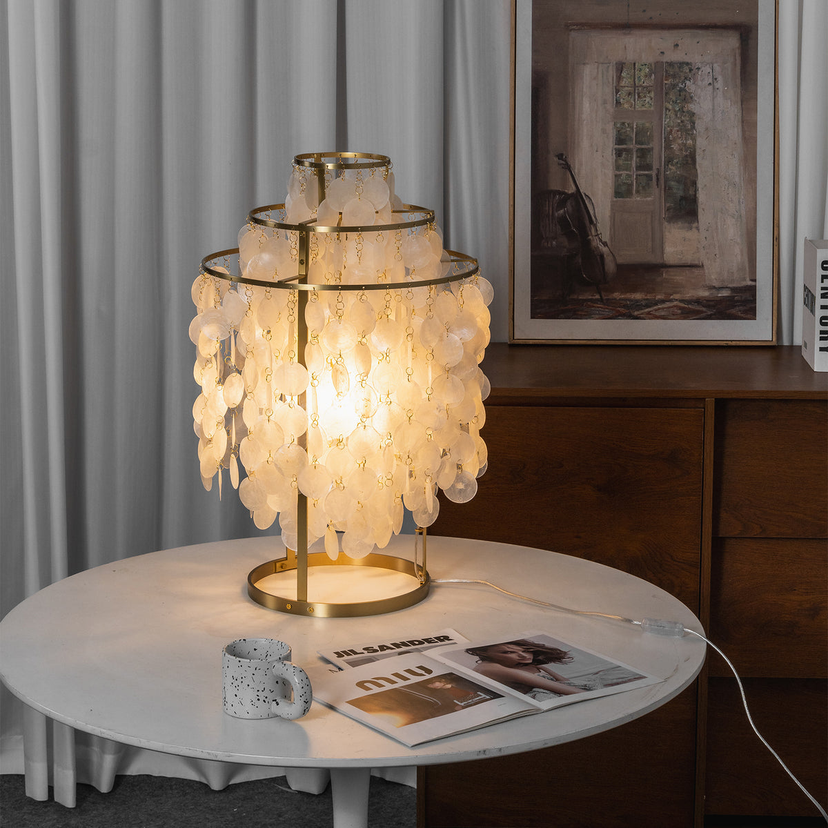 Callista Tiered Capiz Table Lamp | Capiz, Metal | Staged room view of the illuminated seashell lamp casting a warm ambient light alignment on a white pedestal table near a cabinet | Severn Lighting