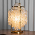 Callista Tiered Capiz Table Lamp | Capiz, Metal | Close-up staged shot of the fully illuminated capiz shell table lamp casting a heavy golden glow onto a polished dark wood table surface | Severn Lighting