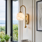 Cordelia Alabaster Globe Wall Sconce | Natural Alabaster and Brushed Brass | Fixed on a wall next to a large glass window showing a scenic view of green forest trees | Severn Lighting