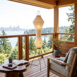 Cosima Hourglass Washi Paper Floor Lamp | Japanese Wabi-Sabi Paper and Black Tripod | Standing on a wooden balcony overlooking scenic forest trees and water | Severn Lighting