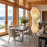 Cosima Hourglass Washi Paper Floor Lamp | Japanese Wabi-Sabi Paper and Black Tripod | Positioned in a modern dining room next to a wooden dining table and large scenic windows | Severn Lighting