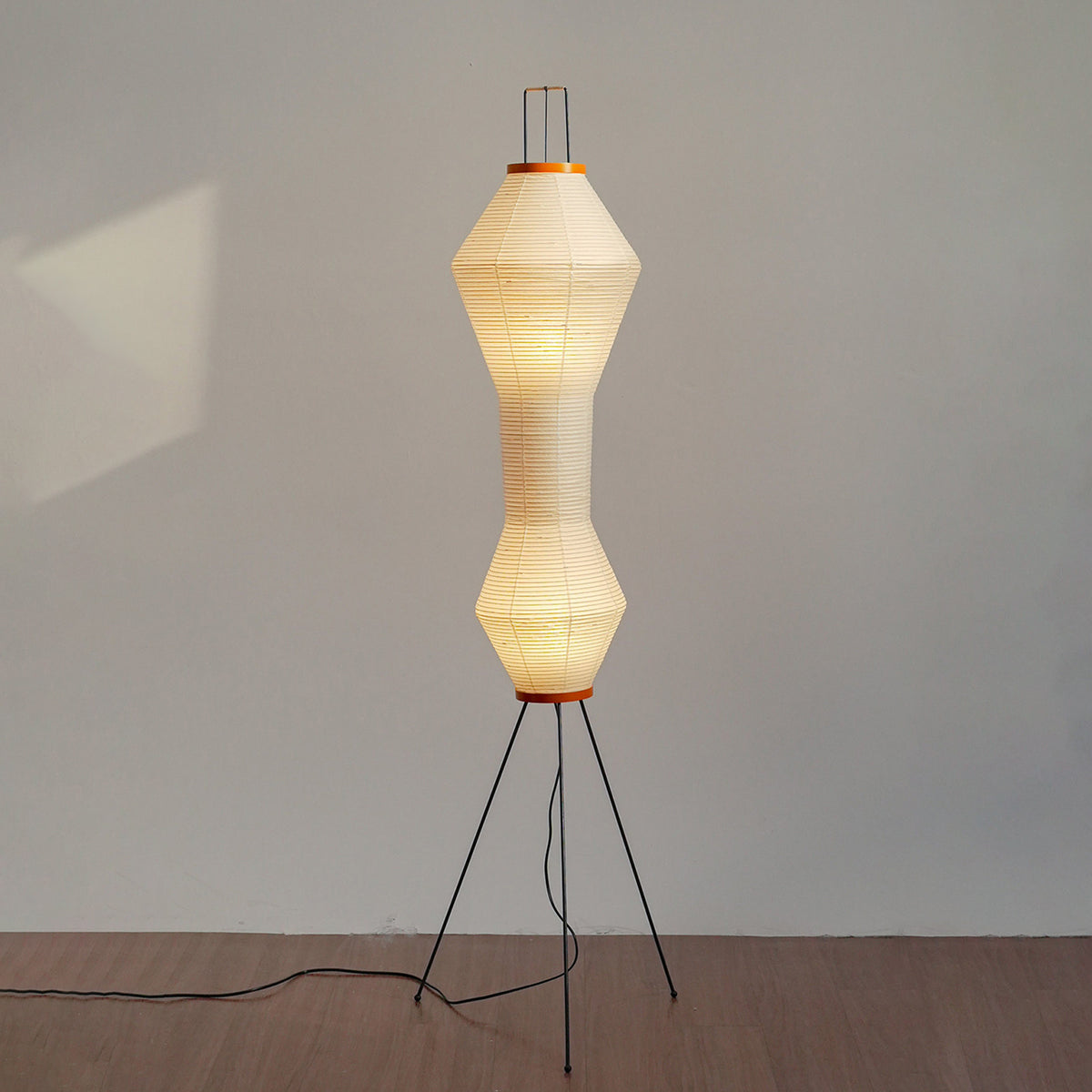 Cosima Hourglass Washi Paper Floor Lamp | Japanese Wabi-Sabi Paper and Black Tripod | Studio view of the hourglass fixture fully illuminated against a plain minimalist wall | Severn Lighting