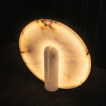 Elvire Alabaster Disc Table Lamp | Natural Alabaster | Angled product view glowing brightly on a dark backdrop | Severn Lighting