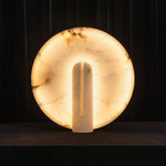 Elvire Alabaster Disc Table Lamp | Natural Alabaster | Front view of the glowing circular stone shade on a dark background | Severn Lighting