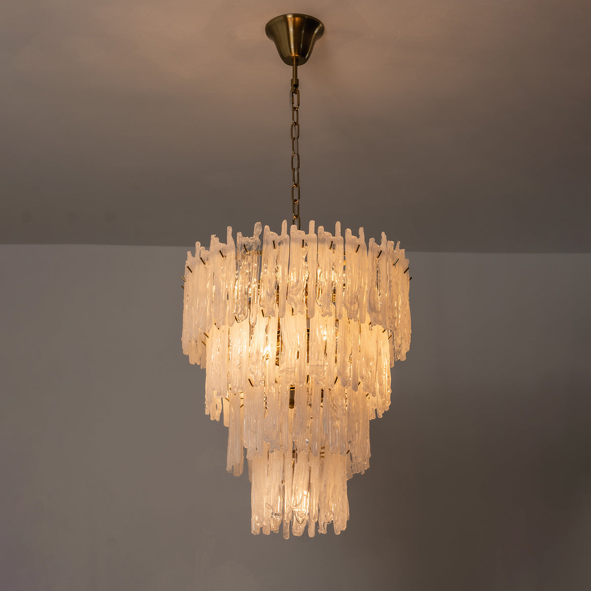 Ethereal Icicle Waterfall Chandelier | Frosted Icicle Glass and Gold Brass | Symmetrical view of the multi-layered fixture fully lit and glowing against a plain dark wall | Severn Lighting