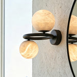 Lucern Twin Globe Alabaster Wall Sconce | Natural Alabaster and Matte Black Metal | Side perspective view showing the fixture glowing next to a round black-framed mirror | Severn Lighting