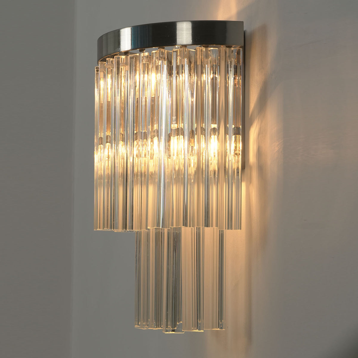 Marelle Glass Rod Tiered Wall Lamp | Clear Glass Rods and Metal | Angled close view of the wall lamp mounted on a neutral wall, glowing softly | Severn Lighting