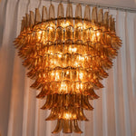 Odile Amber Petal Tier Chandelier | Amber Glass and Metal | Full hanging view glowing warmly against a curtain backdrop | Severn Lighting