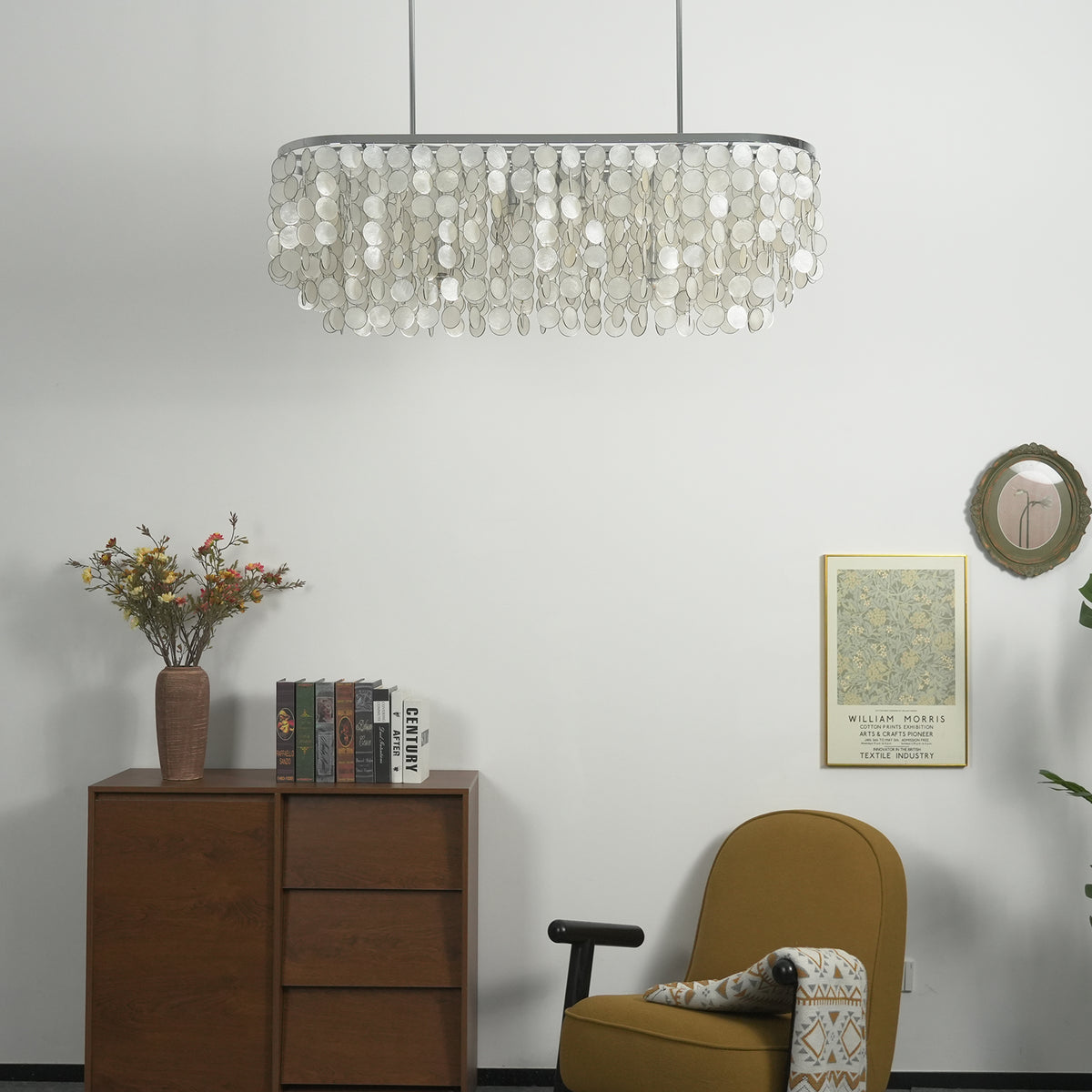 Sirena Capiz Elongated Linear Chandelier | Capiz Shell, Metal | Unlit chandelier hanging in a minimalist corner over a dark wood sideboard cabinet and an accent chair | Samu Lamp Sirena Capiz