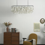 Sirena Capiz Elongated Linear Chandelier | Capiz Shell, Metal | Unlit chandelier hanging in a minimalist corner over a dark wood sideboard cabinet and an accent chair | Samu Lamp Sirena Capiz