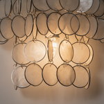 Sirena Capiz Tiered Wall Sconce | Capiz Shell, Metal | Close-up view of the glowing translucent shell circles under a warm yellow internal light | Detail shot | Samu Lamp