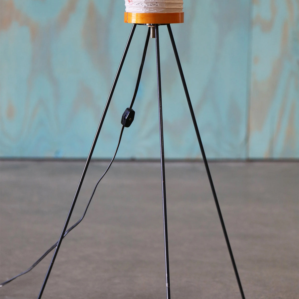 Verena Ribbed Paper Column Floor Lamp | Ribbed Paper and Metal Tripod Legs | Detailed shot focusing on the slender black metal tripod legs and floor switch against a blue wall | Severn Lighting