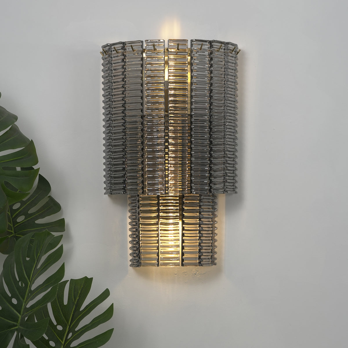 Vespera Tiered Smoky Glass Wall Lamp | Glass, Metal | Studio close-up of the fully illuminated tiered lamp casting a soft warm backlight halo against a neutral grey wall backdrop | Severn Lighting