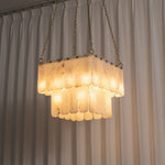 Vivienne Tiered Alabaster Chandelier | Alabaster, Brass | Fully lit tiered square luxury chandelier casting a bright warm gold halo in front of vertical white pleated curtains | Severn Lighting